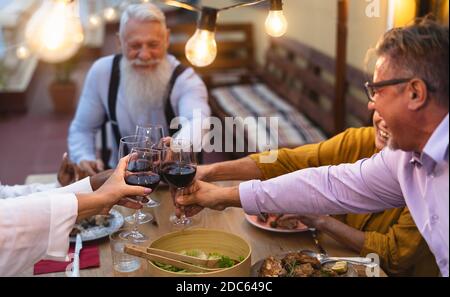 Glückliche multirassische ältere Freunde toasten mit Rotweingläsern zusammen Abendessen auf der Terrasse des Hauses - ältere Menschen und Essen Konzept Stockfoto