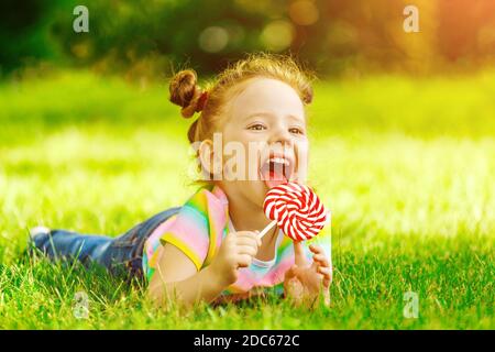 Ein kleines Mädchen mit einem roten Lollipop liegt auf dem Sommerrasen im Park. Stockfoto