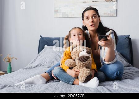 Aufgeregt Mutter und Tochter Fernsehen, während sie auf dem Bett sitzen Mit Teddybär Stockfoto