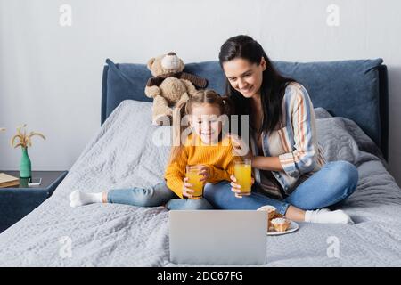 Fröhliche Frau mit Tochter hält Orangensaft während Film Auf einem Laptop Stockfoto