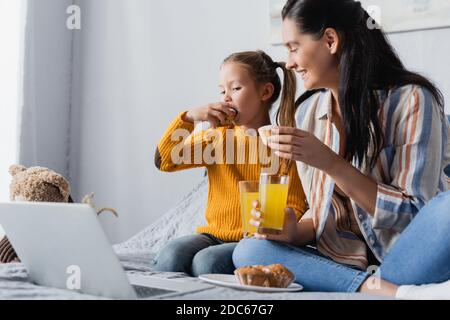 Glückliche Mutter beim Betrachten der Tochter, die Muffin isst, während sie sich einen Film ansieht Auf einem Laptop Stockfoto