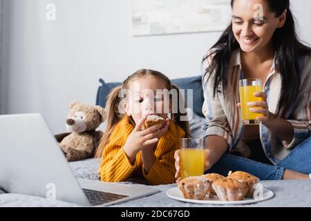 Mädchen essen Muffin in der Nähe fröhliche Mutter, während Film auf Notebook Stockfoto