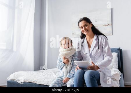 Glücklich Mädchen in Schal und Pyjamas zeigt Daumen nach oben, während In der Nähe des Kinderarztes sitzen Stockfoto