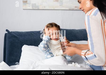 Mutter, die einem kranken Kind ein Glas Wasser gibt Kopfschmerzen beim Sitzen im Bett mit geschlossenen Augen Stockfoto