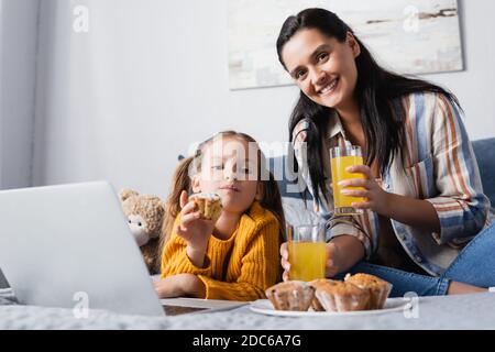 Glückliche Frau und Tochter, die in der Nähe von Muffins und der Kamera schauend Orangensaft beim Ansehen von Filmen auf dem Laptop Stockfoto