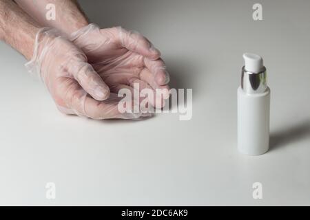 Zwei Hände in halbtransparenten weißen Handschuhen und eine Flasche hydroalkoholisches Gel auf einem weißen Tisch. Covid-19 Schutzausrüstung. Stockfoto