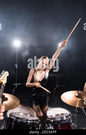 Blonde Frau mit ausgestreckter Hand zwinkert, während sie in der Nähe von Trommel sitzt Kit mit Hintergrundbeleuchtung auf verschwommenem Vordergrund Stockfoto