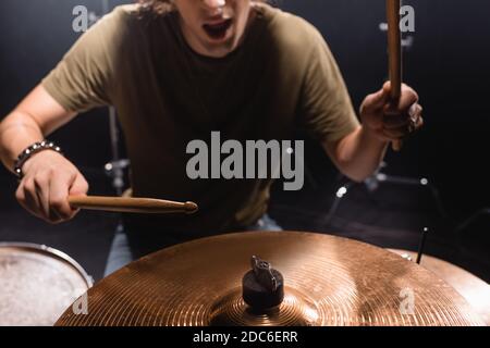 Beschnittene Ansicht des Musikers mit offenem Mund hält Drumsticks und Spielen Becken auf verschwommenem Hintergrund Stockfoto