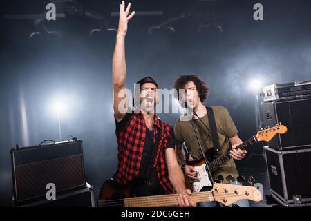 KIEW, UKRAINE - 25. AUGUST 2020: Rockband-Musiker schreien, während sie Gitarren in der Nähe von Mikrofon-Rack und Combo-Verstärker mit Hintergrundbeleuchtung auf Backgrou halten Stockfoto