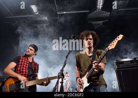 KIEW, UKRAINE - 25. AUGUST 2020: Rockband-Musiker spielen E-Gitarren in der Nähe von Mikrofon-Rack mit Rauch und Frau im Hintergrund Stockfoto