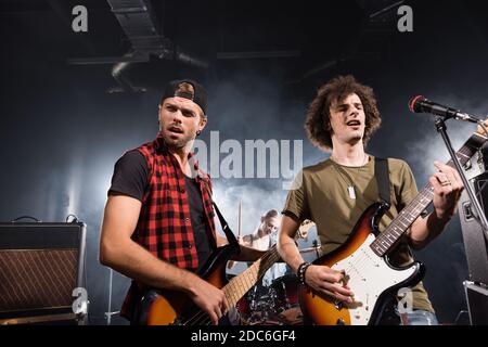 KIEW, UKRAINE - 25. AUGUST 2020: Musiker spielen Bassgitarren und stehen neben einem Mikrofon-Rack und Combo-Verstärkern mit unscharfem Drummer auf dem Hintergrund Stockfoto