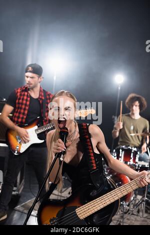 KIEW, UKRAINE - 25. AUGUST 2020: Sängerin der Rockband mit E-Gitarre schreiend im Mikrofon mit hinterleuchteten und verwackelten Musikern auf Backg Stockfoto