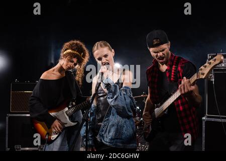 KIEW, UKRAINE - 25. AUGUST 2020: Blonde Sängerin der Rockband singt neben Musikern mit Bass-Gitarren mit Hintergrundbeleuchtung auf schwarz Stockfoto