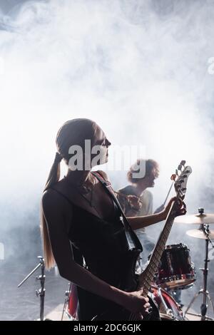 KIEW, UKRAINE - 25. AUGUST 2020: Frau spielt E-Gitarre mit Rauch und verschwommener Schlagzeuger mit Schlagzeug im Hintergrund Stockfoto
