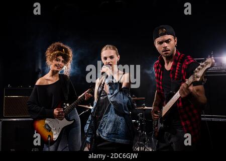 KIEW, UKRAINE - 25. AUGUST 2020: Sängerin der Rockband in der Nähe von Musikern mit E-Gitarren auf schwarz stehen Stockfoto