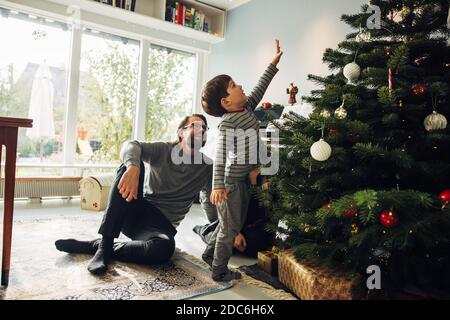 Kind steht am Weihnachtsbaum und reicht seine Hand mit den Eltern beobachten. Kleine Familie zusammen zu Hause während Weihnachten. Stockfoto