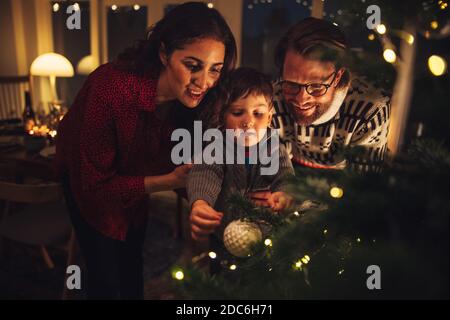 Junge binden dekorative Kugeln auf Weihnachtsbaum mit Eltern zu Hause. Weihnachtsferien Vorbereitungen auf skandinavischen Hause. Stockfoto