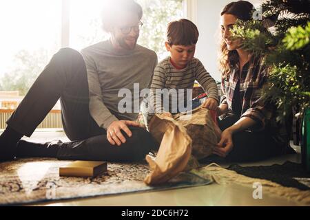 Junge öffnet sein weihnachtsgeschenk mit sitzen mit seinen Eltern am Weihnachtsbaum. Mutter und Vater sehen ihrem Sohn gemütlich zu, wie er sein Weihnachtsgeschenk öffnet. Stockfoto