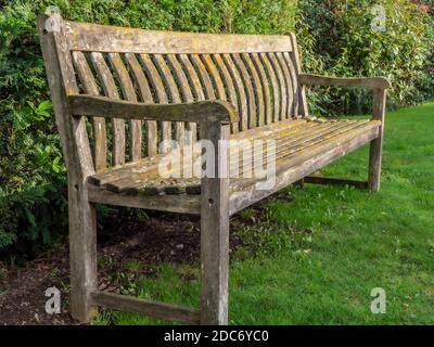 Eine zwanzig Jahre alte, unbesetzte Holzbank / Sitz, mit Blick auf die Sonne, auf Gras, umgeben von grünem Laub. Stockfoto