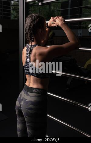 Das Mädchen lehnt sich im Dunkeln an die Sporttreppe. Metallic in einer schönen Form eines Fitness-Studio eines Fitness-Club mit einem Rücken in einem Trainingsanzug für Stockfoto