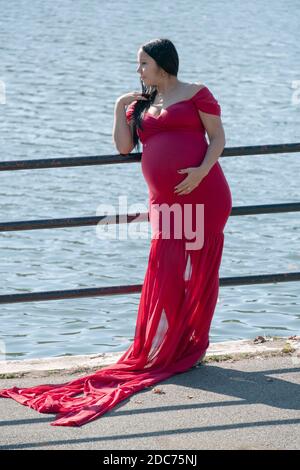 Eine stark schwangere Frau in einem langen fließenden Kleid posiert für Fotos an einem milden Herbsttag. In der Nähe eines Sees in einem Park in Queens, New York. Stockfoto