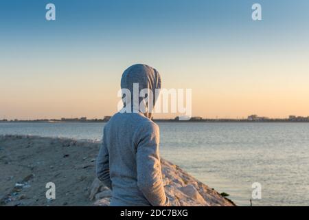 Ein Mann, der eine Jacke mit Hut trägt und dabei beobachtet, wie die Sonne im corniche Park in Dammam, Königreich Saudi-Arabien, aus dem Meer aufgeht Stockfoto