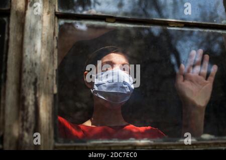 Traurig depressiv ängstlich junge Frau trägt schützende medizinische Maske Blick vor dem Fenster, Coronavirus Covid-19-Virus-Krankheit Pandemie Stockfoto