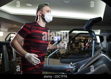 Der junge Mann läuft auf dem Laufband im Fitnessstudio und trägt ein schützendes Gesicht Maske & Latex-Handschuhe aus Gummi, COVID-19 Pandemie Prävention der Ausbreitung & Übertragung Stockfoto