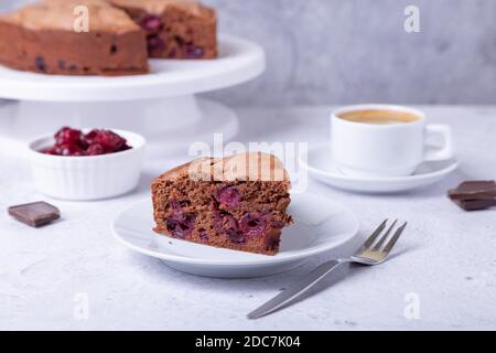 Schokoladenkirschkuchen auf einem weißen Teller. Im Hintergrund ist eine Torte, eine Tasse Kaffee, Kirschen und Schokoladenstücke. Stockfoto