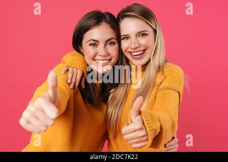 Portrait von zwei glücklichen jungen Freundinnen, die auf rosa Hintergrund stehen und Daumen nach oben zeigen Stockfoto