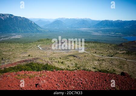 Luftaufnahme zu einem Tal mit geschwungener Straße zu den Bergen. Ausläufer des Osorno Vulkans. Rote vulkanische Felsen im Vordergrund. Osorno, Puerto Varas, Chile Stockfoto