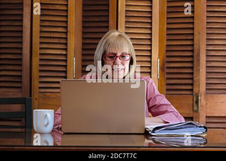 Ältere Frau, die mit einem Laptop arbeitet und Papiere sucht. Sie ist drinnen in einem Holztisch mit einem silbernen Computer. Vorderansicht. Stockfoto