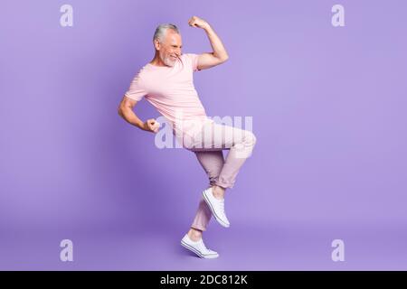 In voller Länge Profil Foto von pensionierten Mann heben Faust Knie Palmen tragen rosa T-Shirt Hose Sneakers isoliert violette Farbe Hintergrund Stockfoto