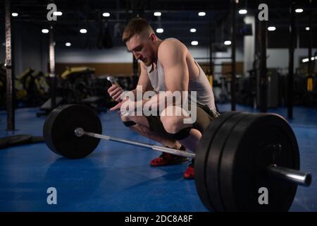 Männlicher Athlet sitzt auf Hacken in der Nähe von Langhantel während der Vorbereitung Heben Sie das Gewicht an Stockfoto