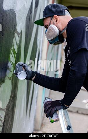 Graffiti-Künstler in Aktion, Zeichnung an der Wand mit Aerosol-Sprühfarbe in einer Dose, tragen Schutzmaske / Atemschutzmaske mit Filtern. Stockfoto