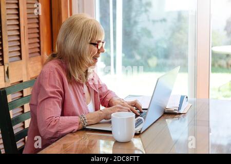 Lustige ältere Frau mit Laptop. Lächelnde Großmutter auf einem Holztisch mit einem silbernen Computer. Kaffeetasse beiseite. Stockfoto