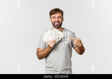 Porträt von schönen bärtigen männlichen Modell in grauem T-Shirt, zeigt auf Geld und lächelt, bekommen einen Kredit, stand über weißem Hintergrund Stockfoto