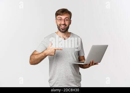 Bild von gutaussehenden Mann mit Bart, trägt Brille und graues T-Shirt, zeigt auf Laptop und lächelt, empfehlen etwas, über weiß stehen Stockfoto