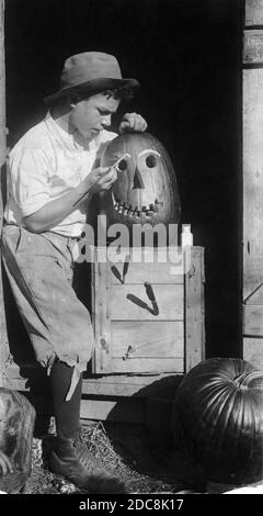 In den 1910er Jahren hat ein Landjunge einen großen Kürbis in eine Jack-o-Lantern mit einem großen, toothy Lächeln geschnitzt. Er konzentriert sich nun darauf, dem Kürbis weiße Augenbrauen ins Gesicht zu malen. Die Kürbiskiste hat zwei geöffnete Jackmesser, die in sie kleben, und zusammen mit dem Jungen, ist es alles in einem Schuppen Tür gerahmt. Unten rechts wartet ein zweiter Kürbis unter dem Messer auf seine Wendung. Um meine anderen Urlaubsbilder zu sehen -- Suche: Prestor vintage Holiday Stockfoto