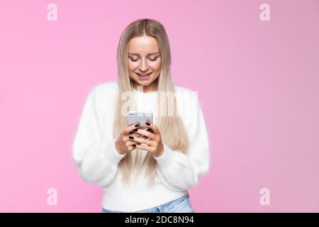 Fröhliche junge Frau lächelnd und mit Handy, Eingabe sms, Nachricht, in flauschigen weißen Pullover gekleidet, isoliert über Studio rosa Hintergrund Stockfoto