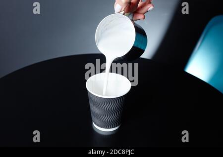 Der Barista gießt Milch in den Einweg-Papierbecher auf dem Holztisch im Café. Zubereitung von leckeren Kaffee trinken mit Milch. Frau hält den Krug mit Metallmilch in der Hand Stockfoto
