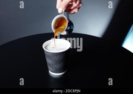 Der Barista gießt Espresso in die Einweg-Papiertasse auf dem Holztisch im Café. Zubereitung von leckeren Kaffee trinken mit Milch Stockfoto