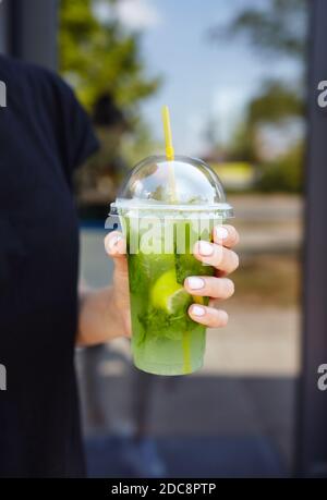 Weibliche Hand hält Plastikbecher mit einem Mojito in einem Stadtcafe. Frisches Cocktailgetränk mit Eisfrucht- und Kräuterdeko. Stockfoto