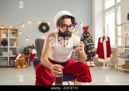 Junger Mann in Unterhemd und Weihnachtsmütze auf Stuhl sitzend Zu Hause am Weihnachtstag und trinken Whisky Stockfoto