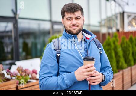 Junger Geschäftsmann mit einer Tasse Kaffee zum Mitnehmen. Bärtiger Mann in blauer Jacke trinkt Cappuccino im Freien. Stockfoto