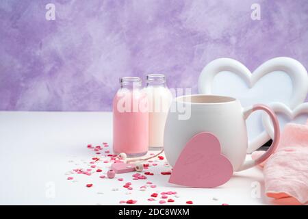 Romantischer Hintergrund mit einem rosa Herz auf einem Kaffeebecher, weißem Rahmen, Milch und Herzen auf einem weißen und violetten Hintergrund. Stockfoto