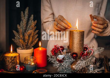 Frau Hände Blitz große goldene Kerze mit Streichhölzern in Weihnachtskranz. Weihnachten Innendekoration auf rustikalem Holzhintergrund. Hygge, Ornamente, Kerze Stockfoto