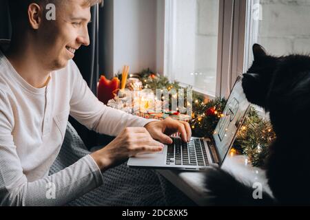 Junger Mann Freiberufler in Pyjama Arbeitsabstand online mit Videoanruf auf Laptop-Computer mit Katze zu Hause im Büro. Weihnachtsstimmung, Neujahr decorati Stockfoto
