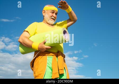 Gesundheitswesen Fröhlicher Lebensstil. Großvater Sportler auf blauen Himmel Hintergründe. Älterer Mann im Health Club. Älterer Mann mit einer Yogamatte. Stockfoto