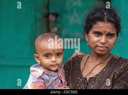 Chikkanayakanahalli, Karnataka, Indien - 3. November 2013: Nahaufnahme Porträt der stolzen jungen Mutter mit Kleinkind auf dem Arm. Grün verblasste Hintergrund. Stockfoto